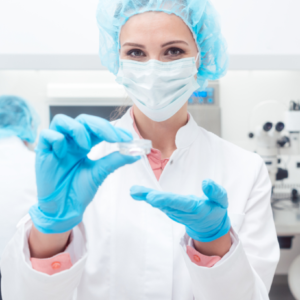 Médica com jaleco branco, touca azul e luvas azuis segurando um pequeno recipiente utilizado no congelamento de óvulos.