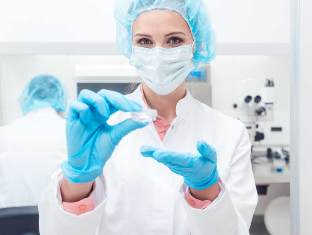 Médica com jaleco branco, touca azul e luvas azuis segurando um pequeno recipiente utilizado no congelamento de óvulos.