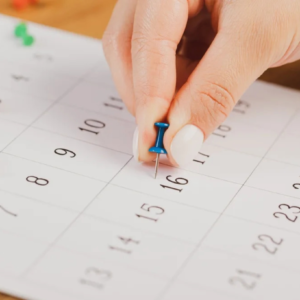 Mão posicionada em um calendário, com um alfinete tipo taça em mãos, remetendo a algum tipo de planejamento, podendo ser o coito programado.