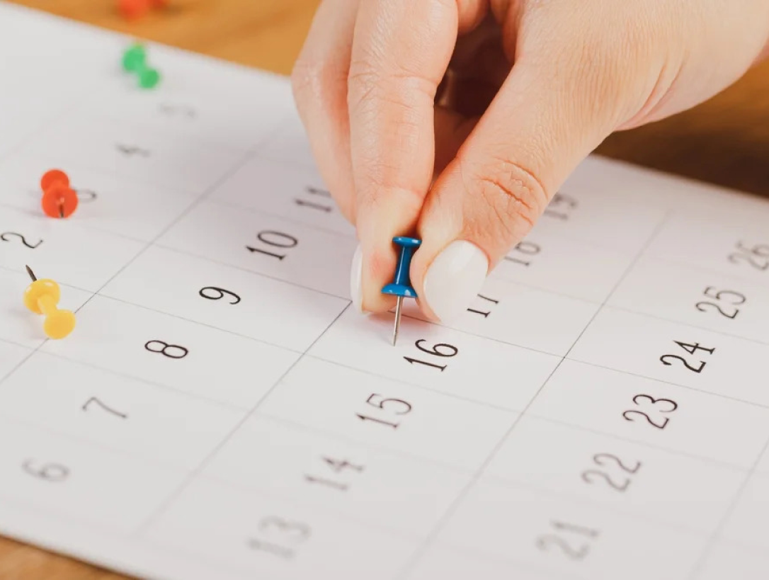 Mão posicionada em um calendário, com um alfinete tipo taça em mãos, remetendo a algum tipo de planejamento, podendo ser o coito programado.