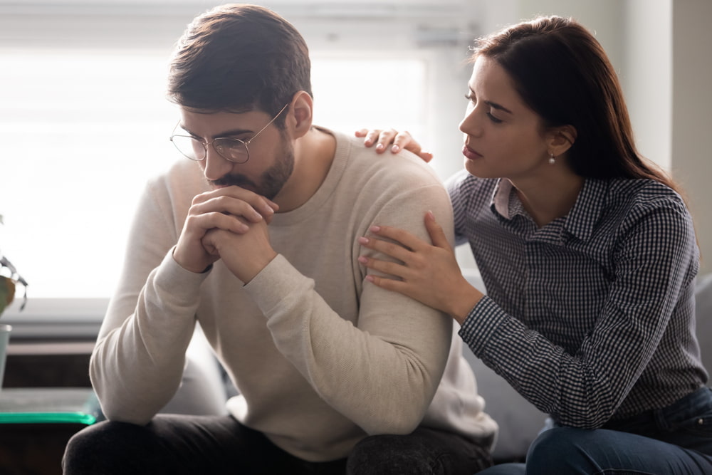 Casal hétero jovem e branco. O homem está frustrado e a mulher tentando consolá-lo.