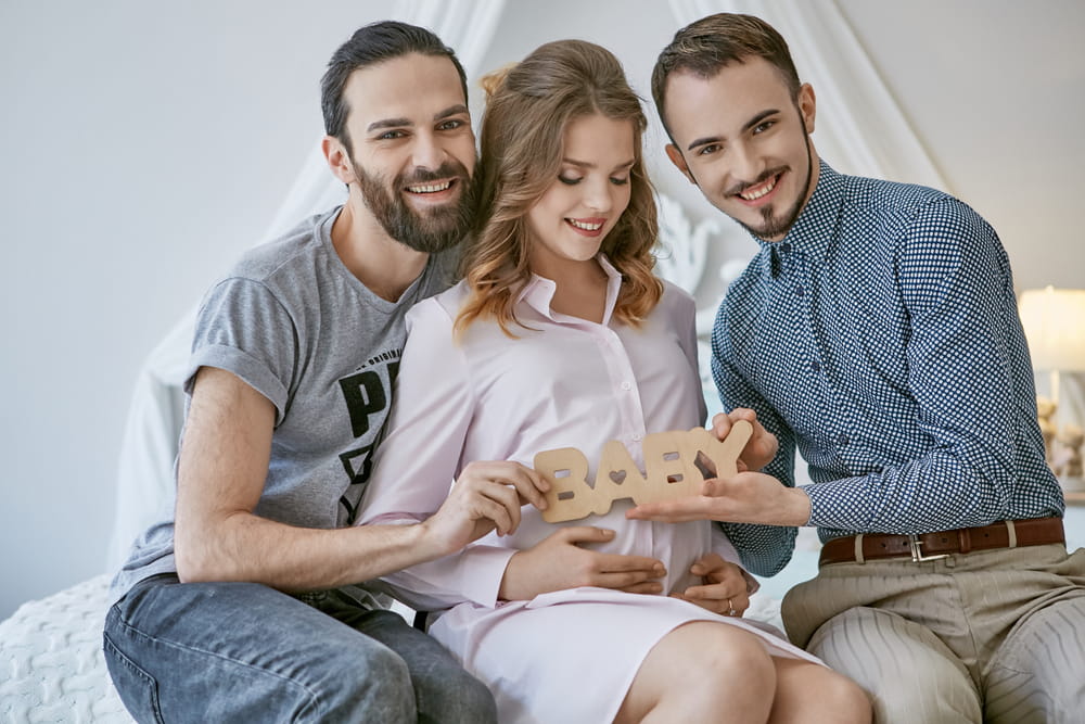 Casal homoafetivo masculina e uma mulher grávida no meio dos dois. Todos com expressão de felicidade, e os dois homens do casal estão segurando um letreiro de madeira escrito "baby", sugerindo que um útero de substituição.